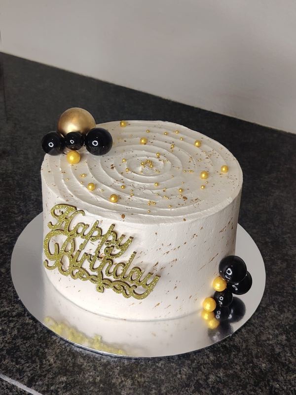 White birthday cake with gold swirl piping, gold Happy Birthday topper and black spheres